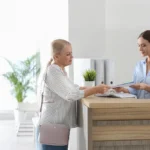 A receptionist checking a woman into a healthcare facility at the counter, with her doctor in the background.